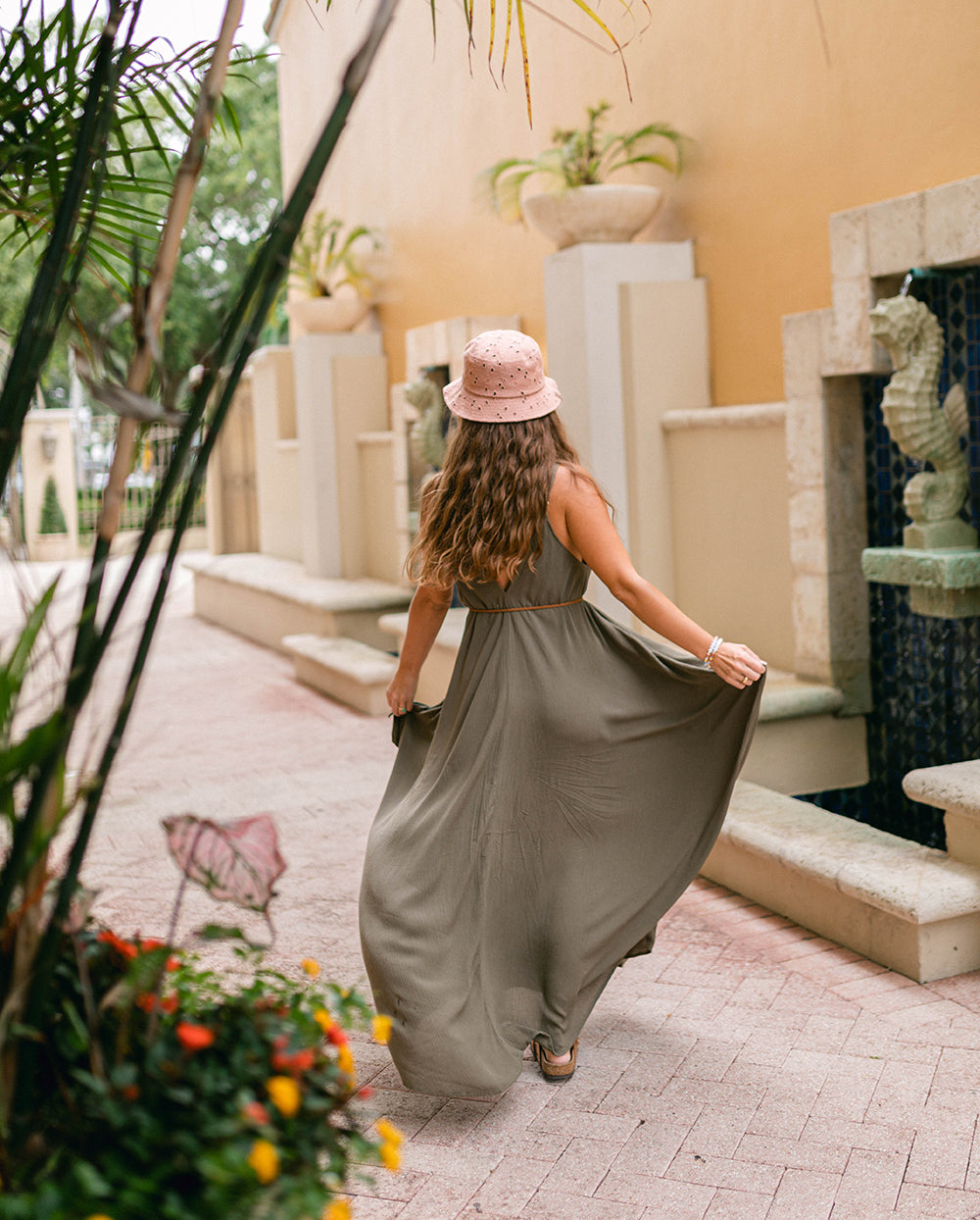 Woman in a long dress and pink hat walking outdoors near plants and a building. #color_basil