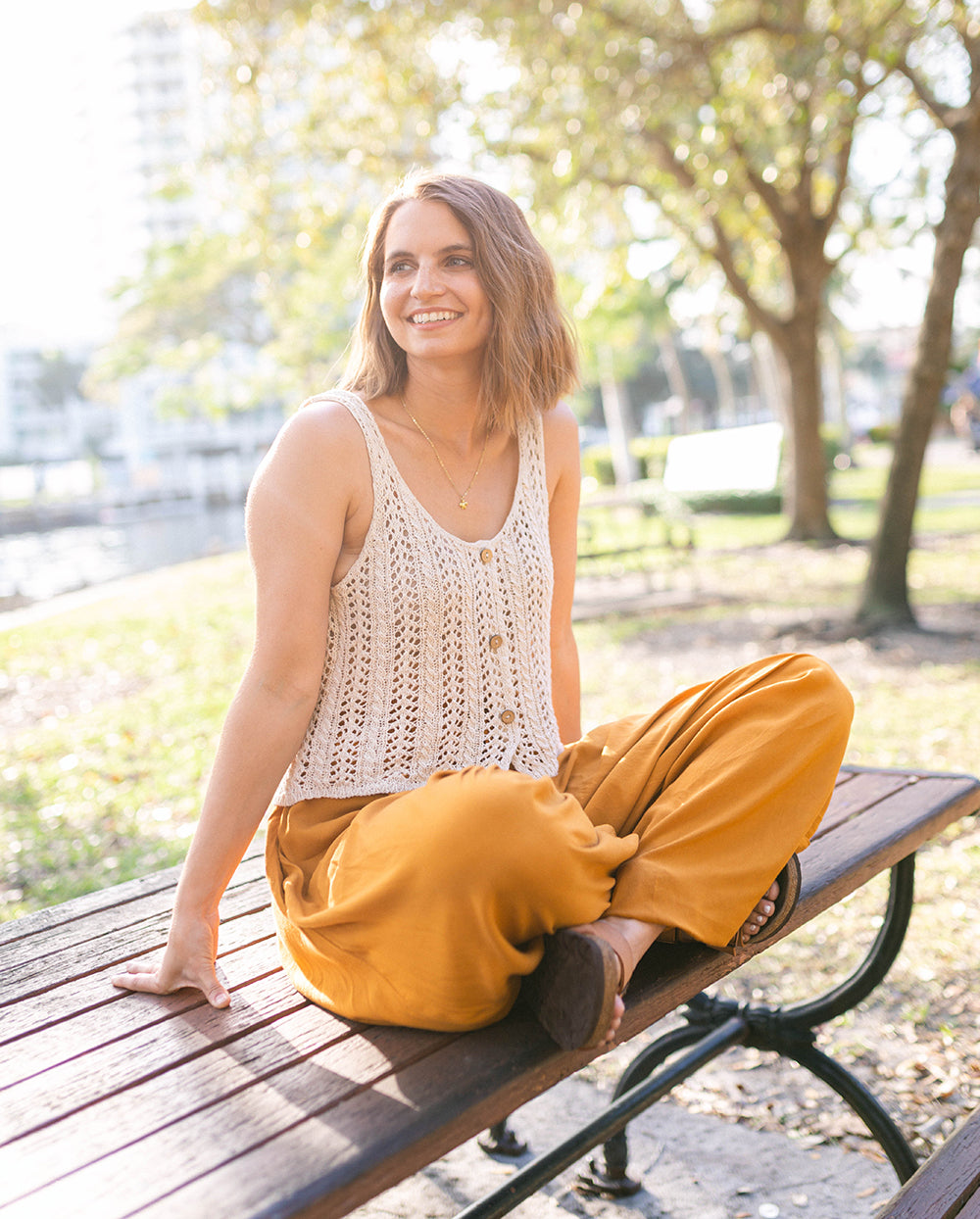Woman sitting on a park bench wearing a white sleeveless top and yellow pants. #color_amber