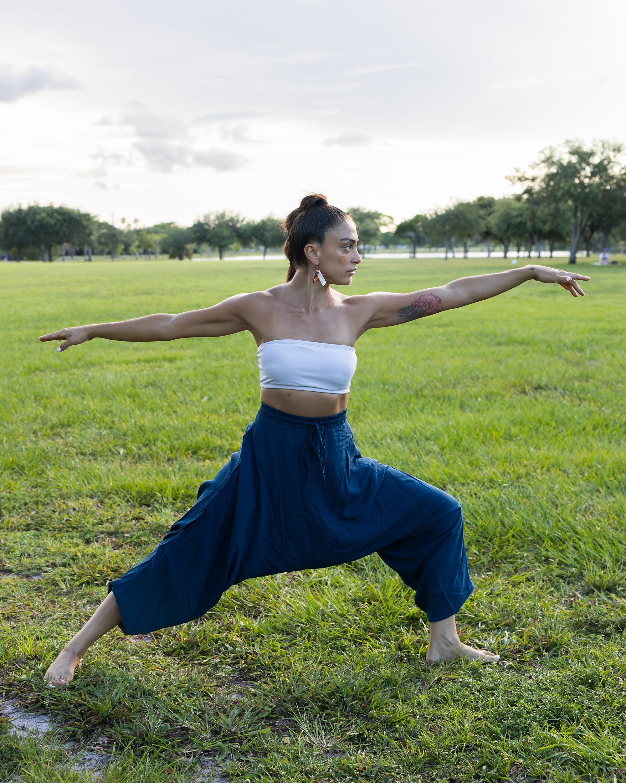 A woman in a yoga warrior pose on a grassy park field, wearing a white strapless tube top and loose navy blue harem pants. #color_marine