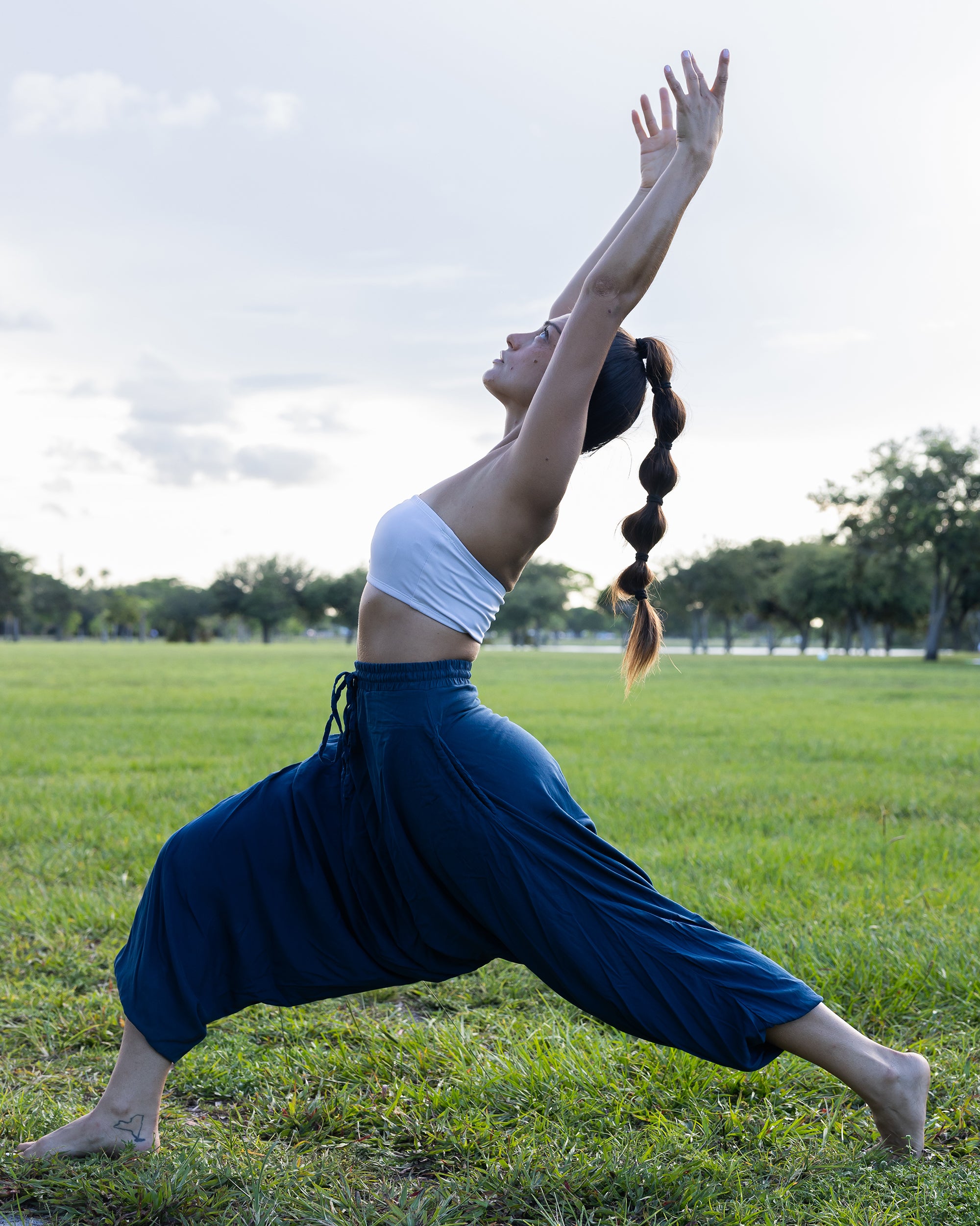 A woman in a yoga pose outdoors on a grassy field, wearing loose navy blue harem-style pants and a white crop top, with trees and a cloudy sky in the background. #color_marine