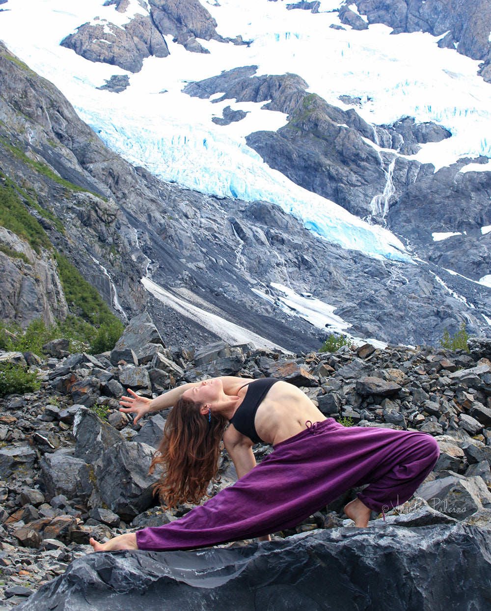 Person in purple pants and black top performing a yoga pose on rocky terrain with a glacier in the background #color_purple
