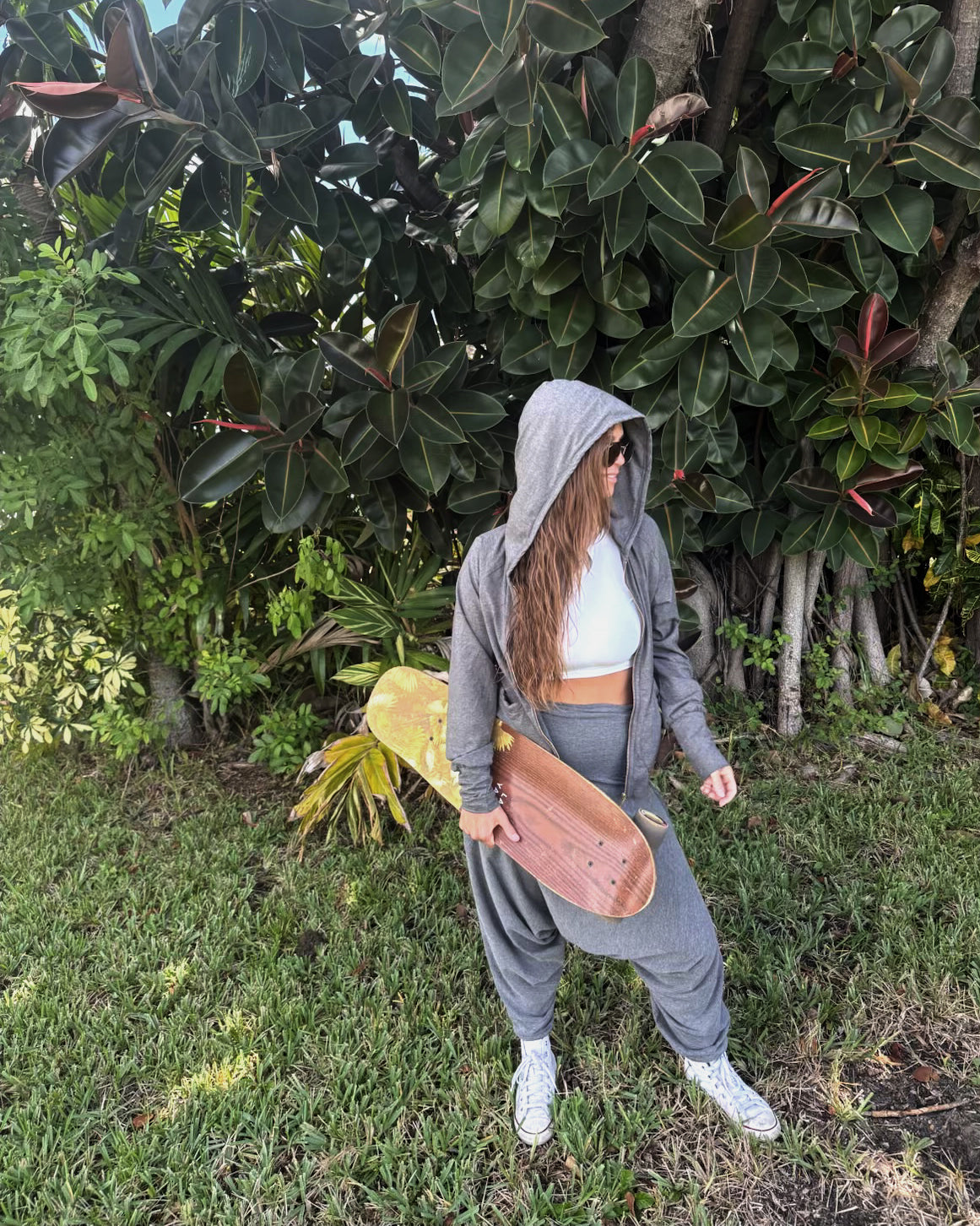 Person in gray hoodie and pants holding a pink surfboard in front of green foliage #color_grey