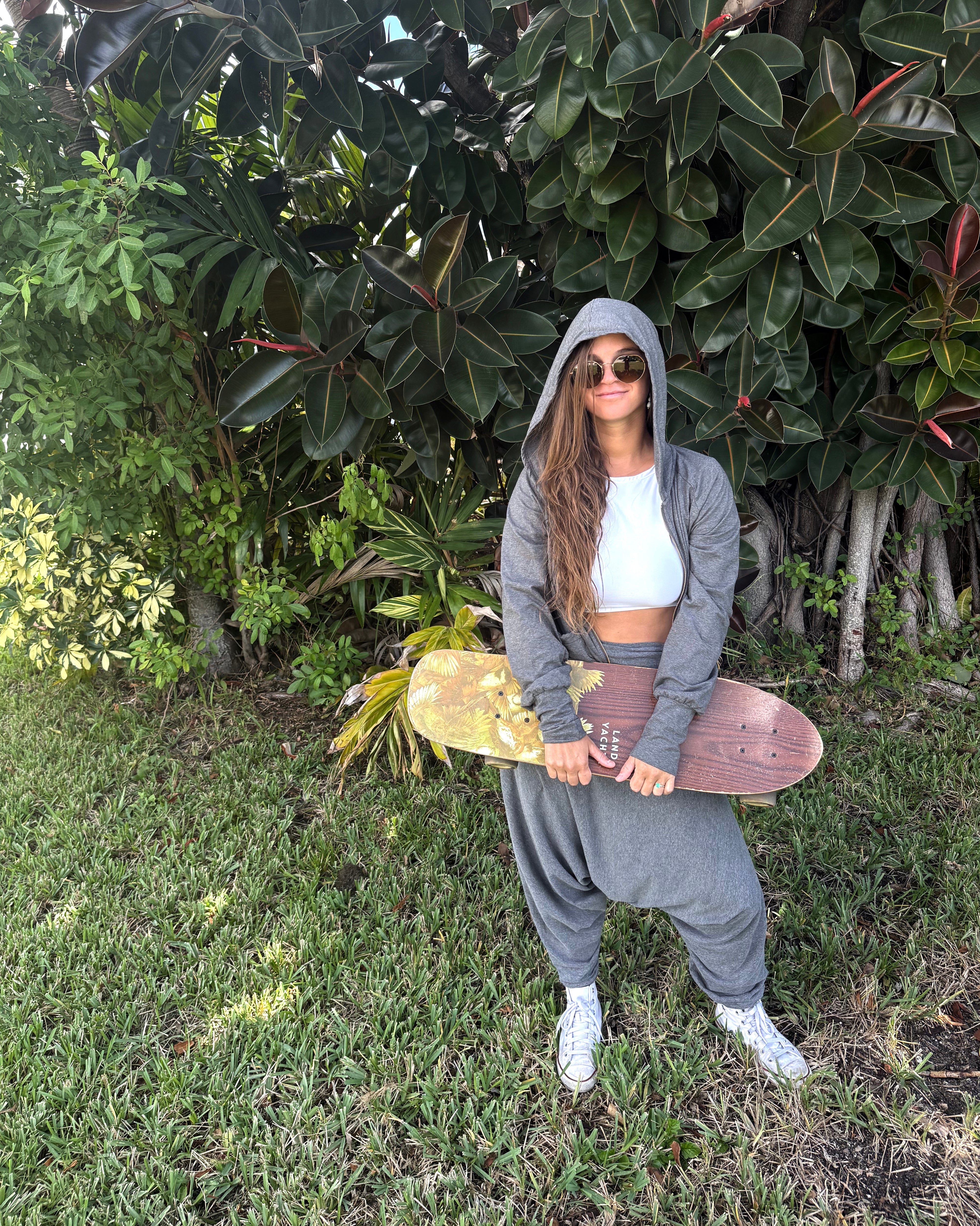 Person holding a skateboard in a grassy area with greenery and harem pants #color_grey