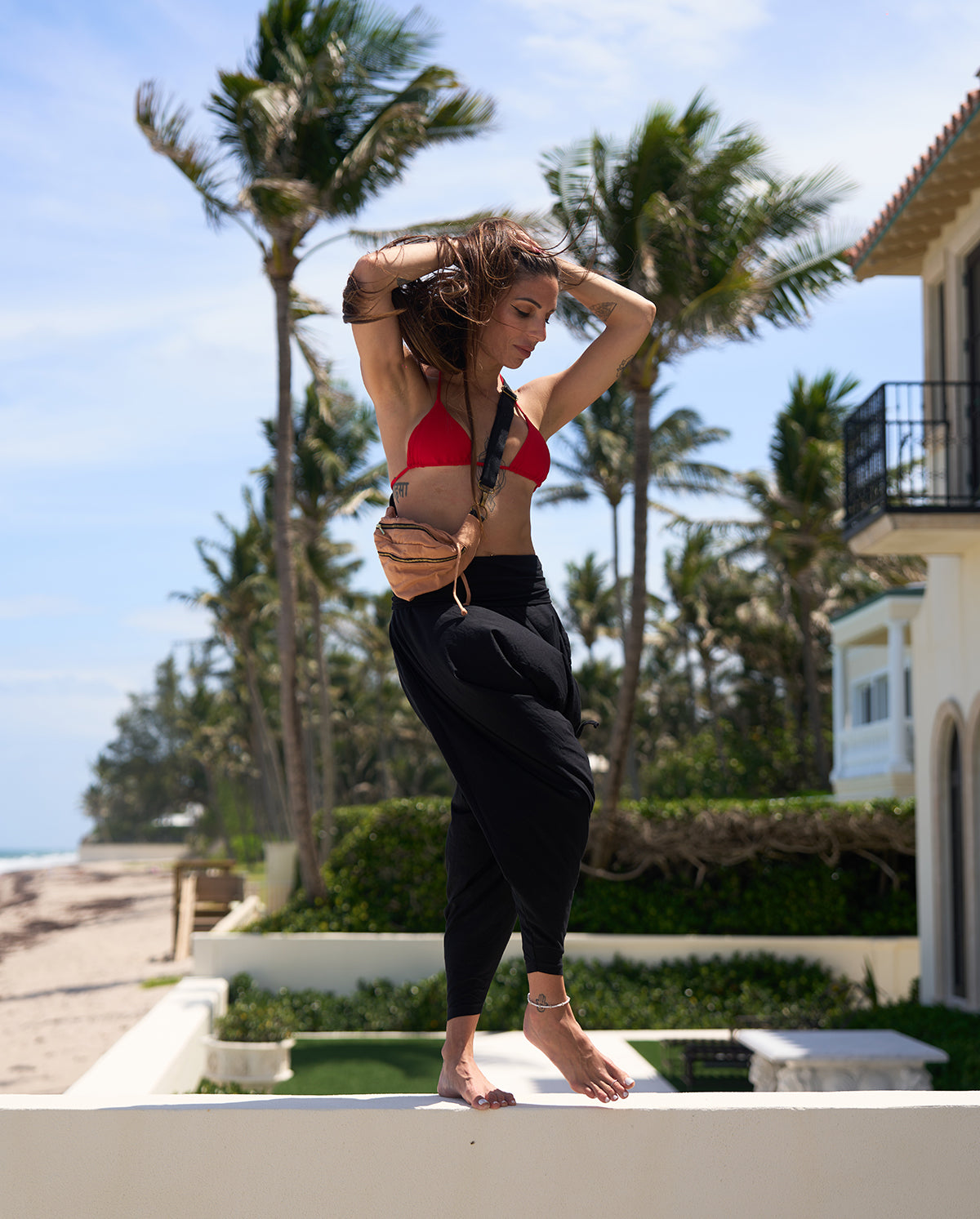 Woman in a red bikini top and black pants posing outdoors with palm trees and a building in the background. #color_camel