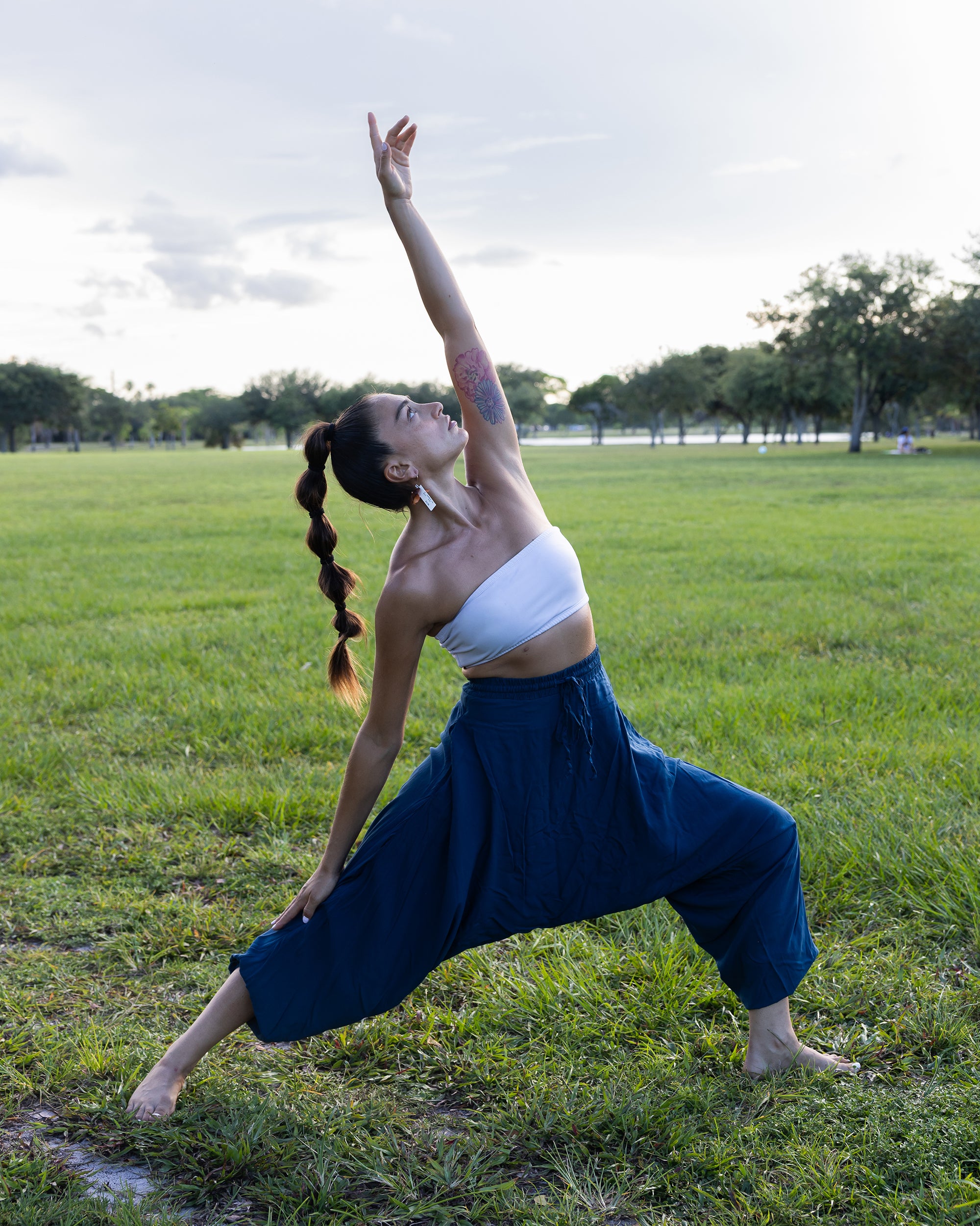 A woman in a yoga pose outdoors on a grassy field, wearing a white tube top and loose dark blue pants, with trees and a cloudy sky in the background. #color_marine