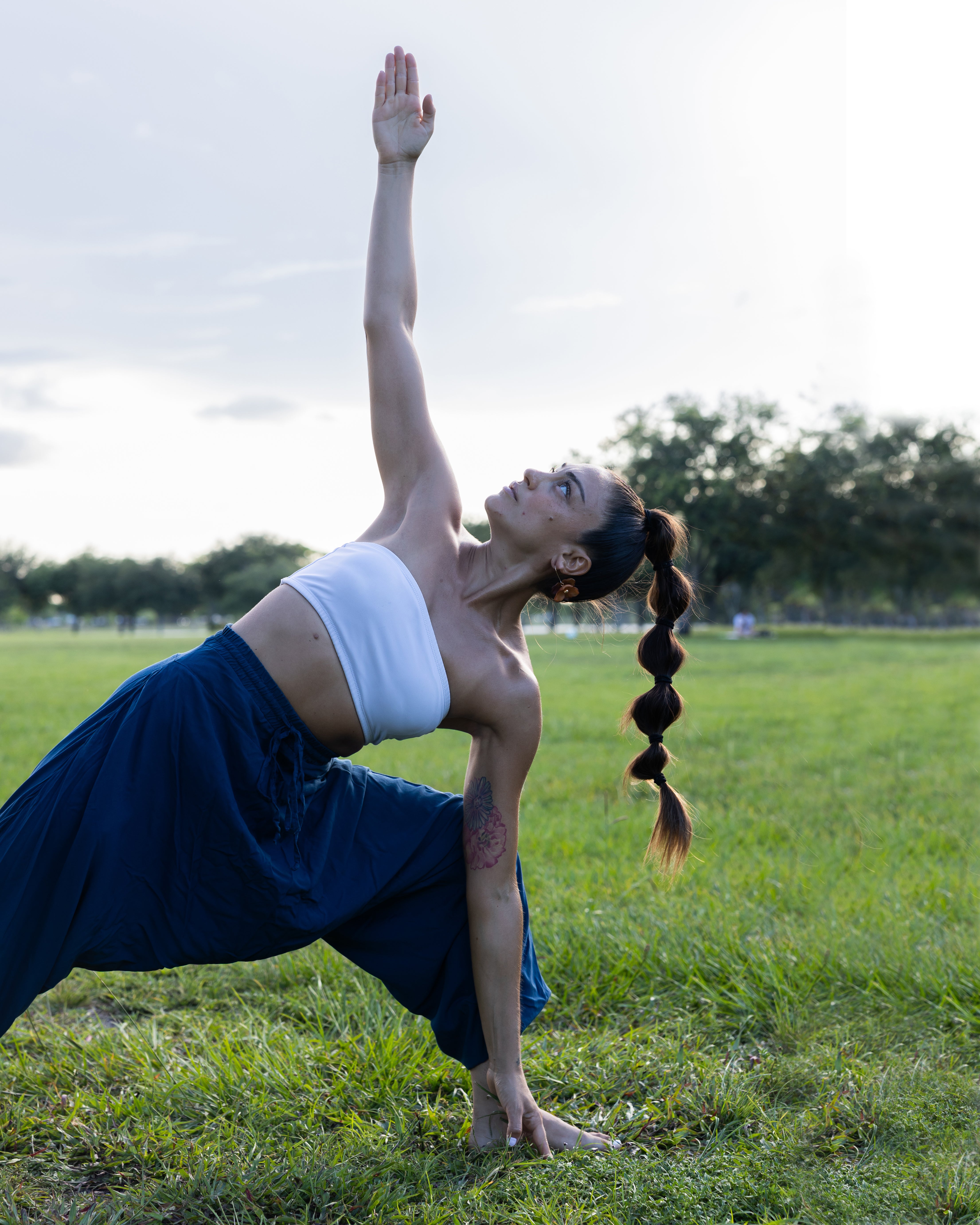A woman in a yoga pose outdoors on a grassy field, wearing a white tube top and a loose dark blue skirt. Trees and a bright sky are visible in the background.#color_marine