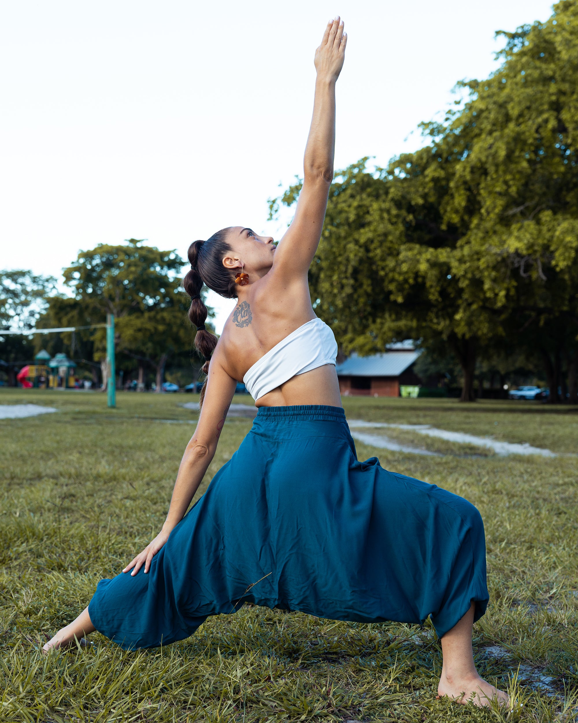 A woman in a yoga pose outdoors on a grassy field, wearing a white wrap-style top and a loose teal-blue wide-leg skirt. Trees and a park are visible in the background. #color_marine