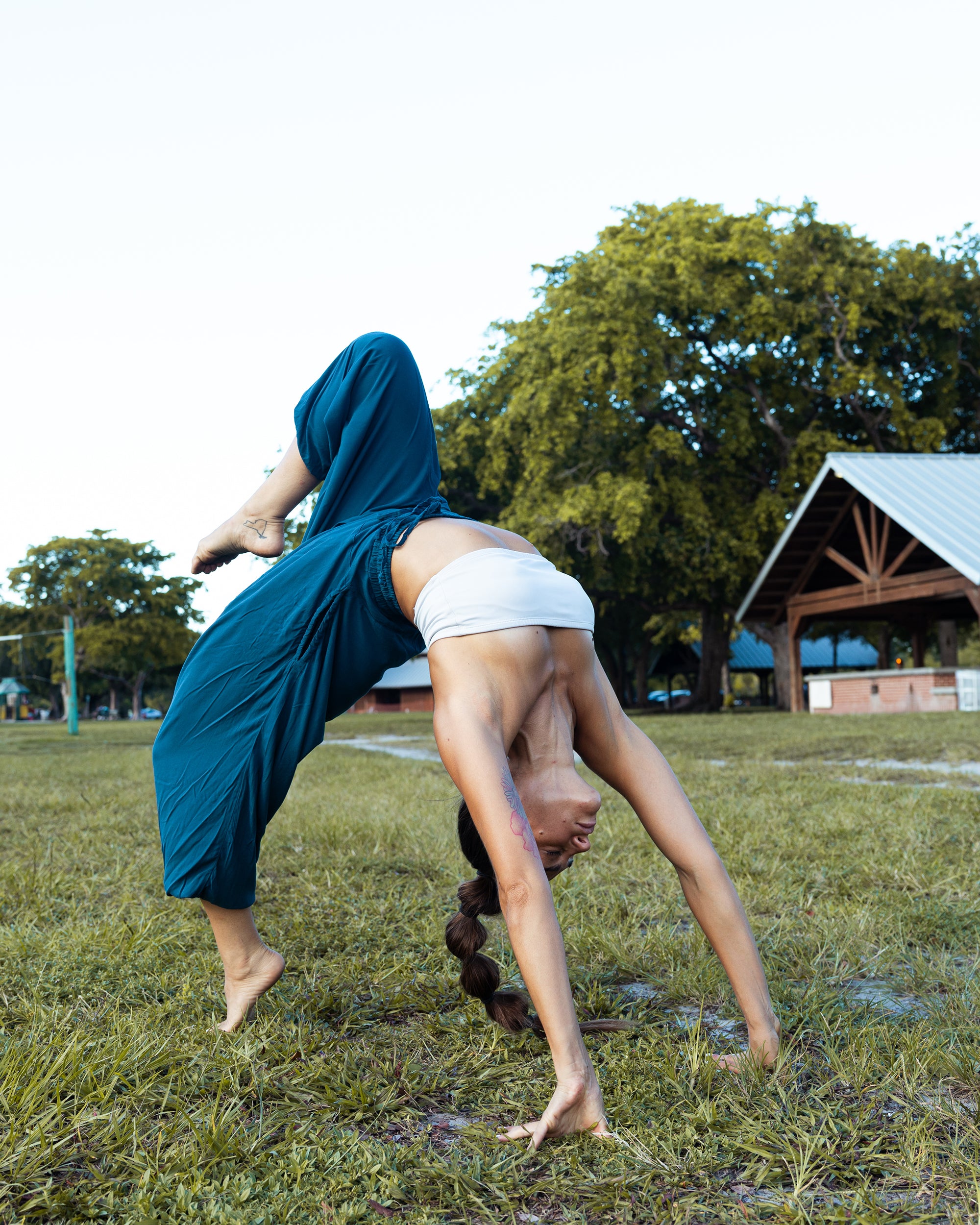 A woman performing a backbend yoga pose on a grassy field, wearing loose teal pants and a white sports bra, with trees and a pavilion in the background. #color_marine