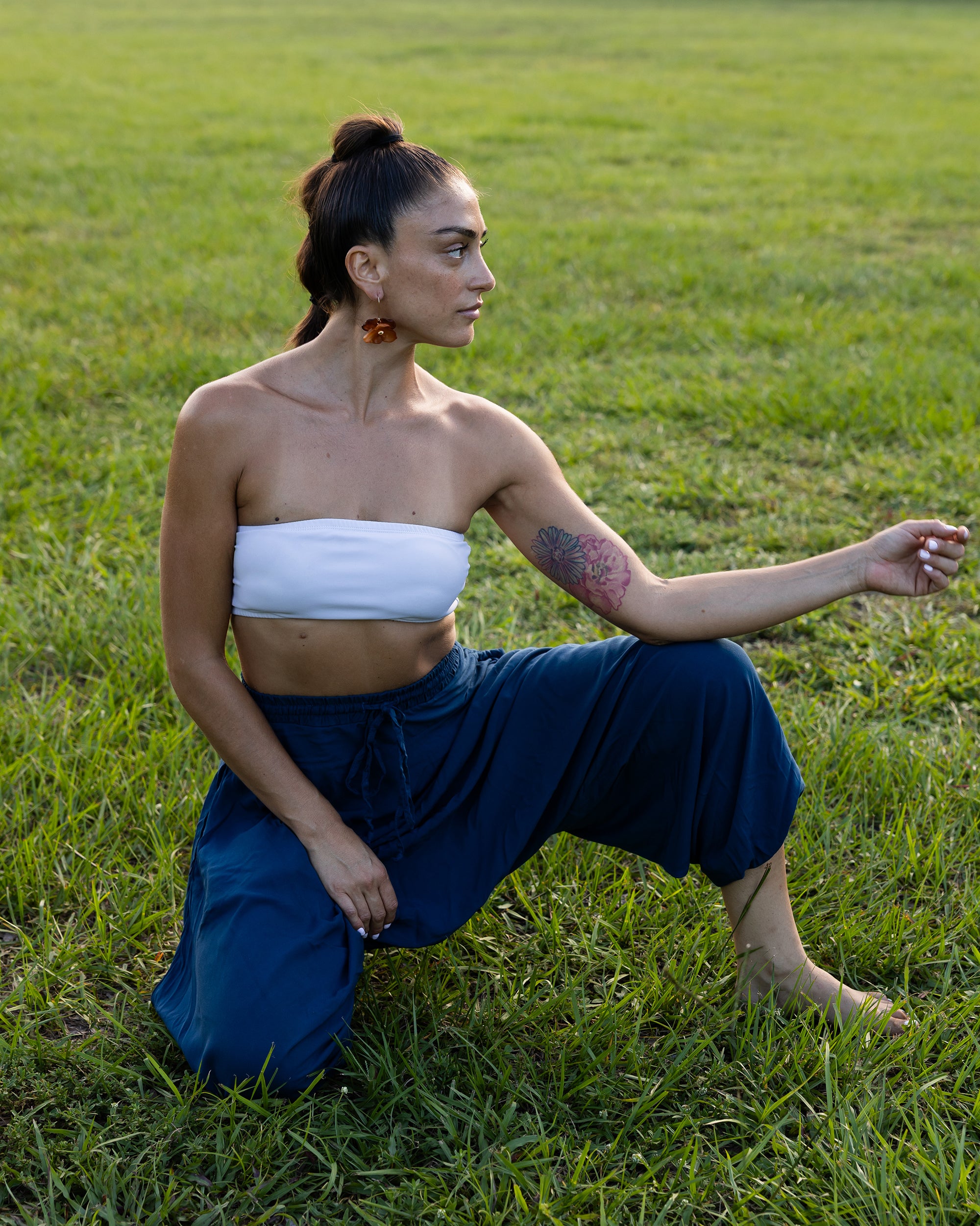 A woman kneeling in a grassy field wearing a white strapless bandeau top and dark blue wide-leg pants. #color_marine