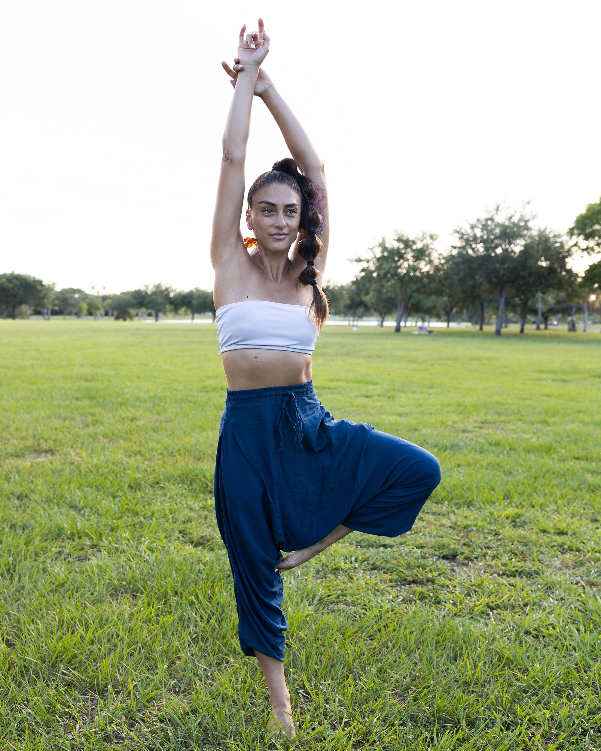 A woman in a yoga tree pose outdoors on a grassy field, wearing a white strapless tube top and dark blue harem-style pants.#color_marine