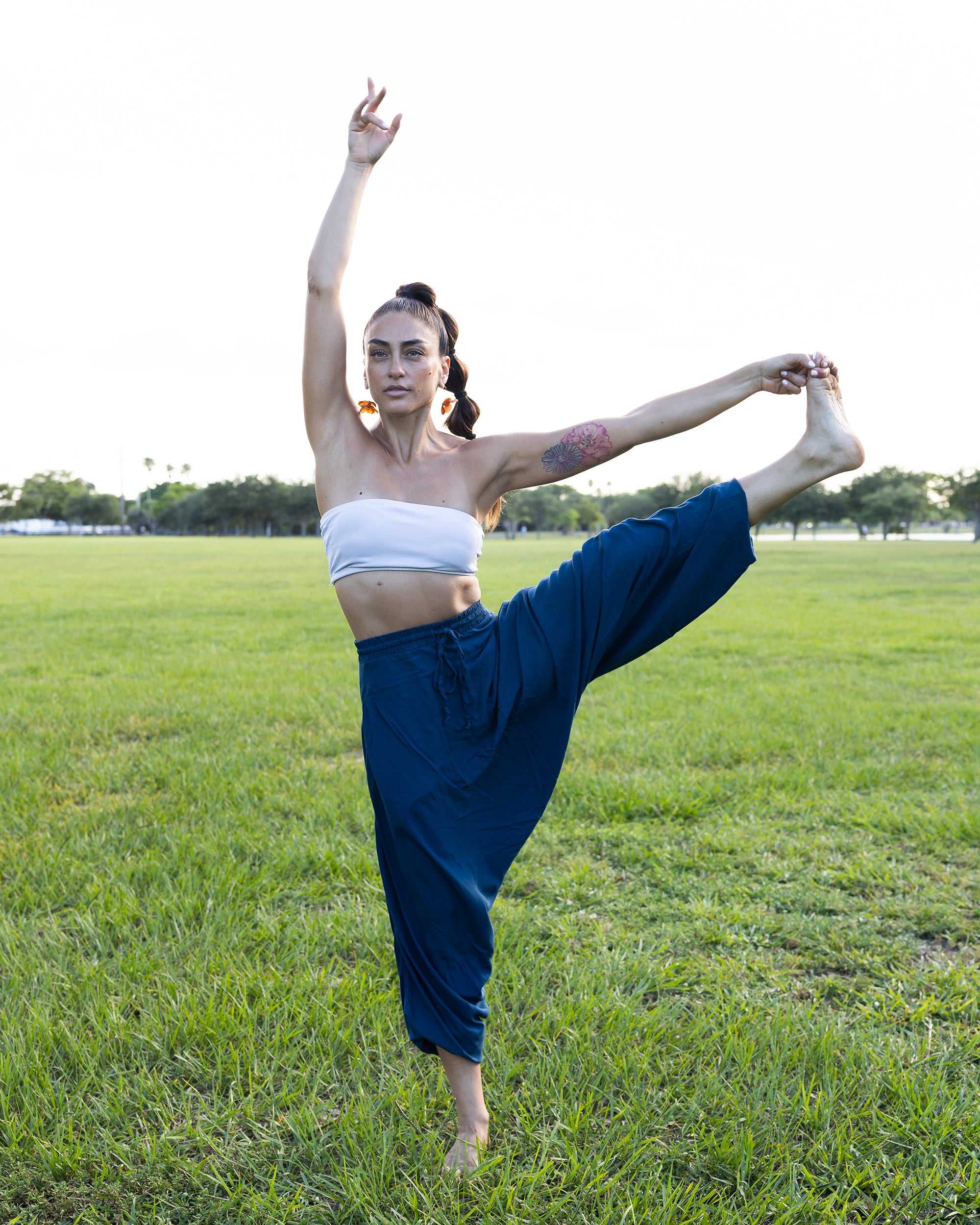 A woman in a yoga pose outdoors on a grassy field, wearing a white bandeau top and loose navy blue pants. #color_marine