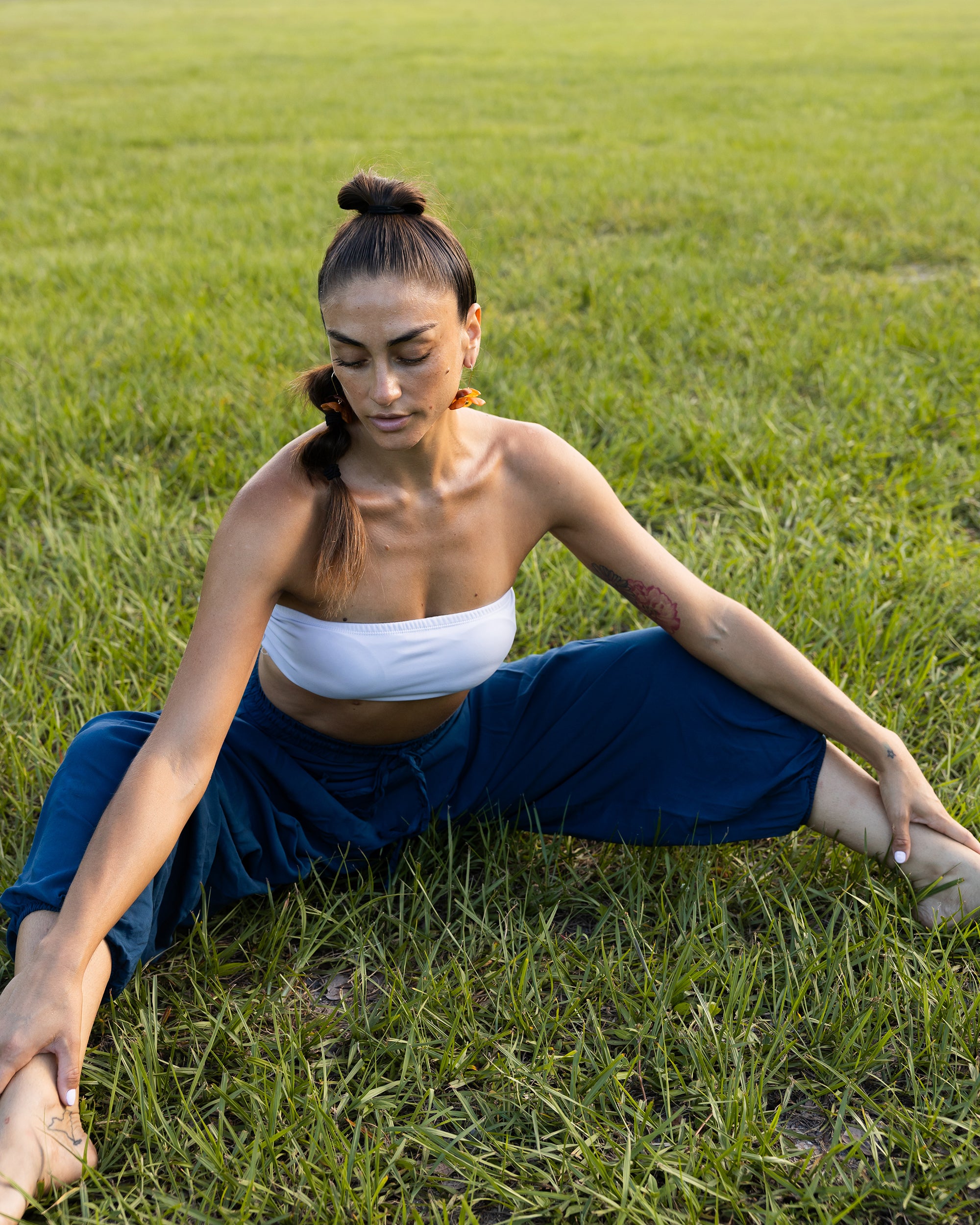 A woman in a white strapless top and navy blue pants sits in a grassy field, stretching her legs wide apart. #color_marine