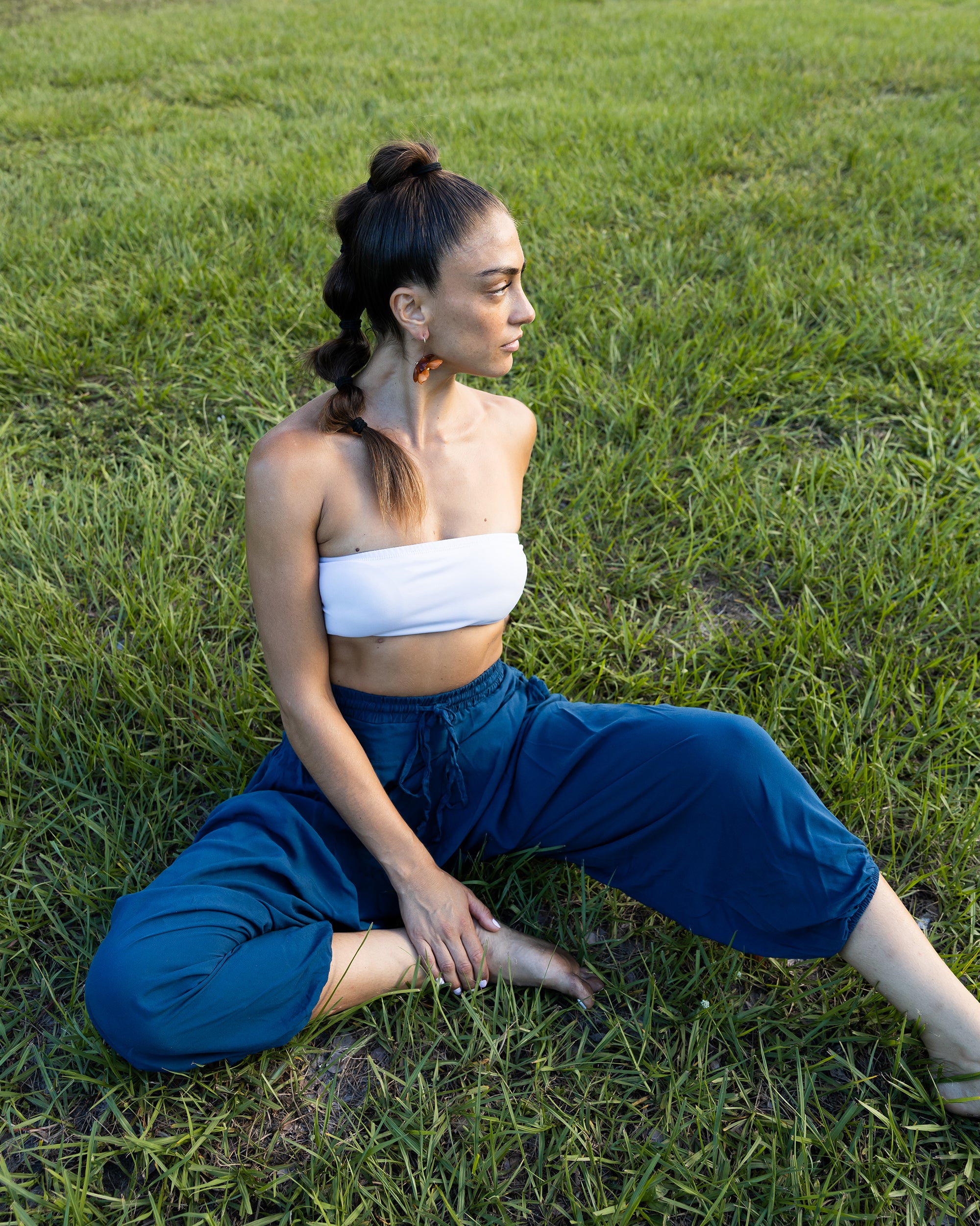 A woman sitting cross-legged on a green grass lawn, wearing a white strapless bandeau top and dark blue cropped pants.#color_marine