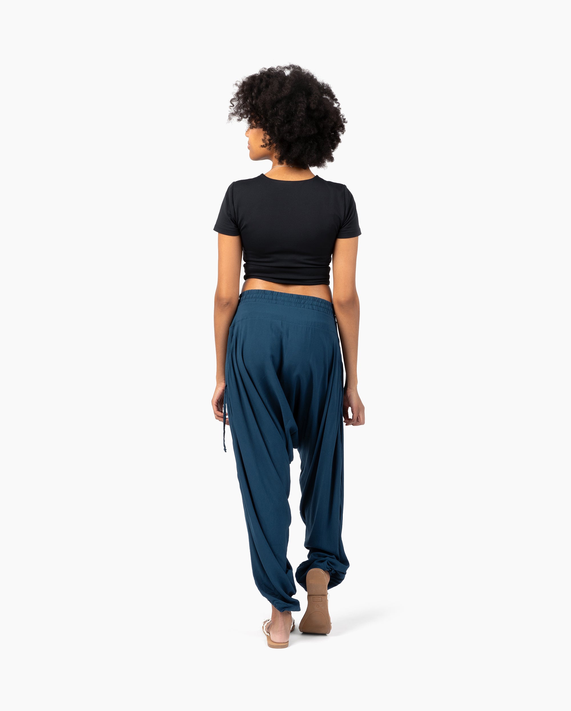 A woman with curly black hair is shown from behind wearing a black short-sleeve crop top and dark teal blue harem pants against a plain white background. #color_marine