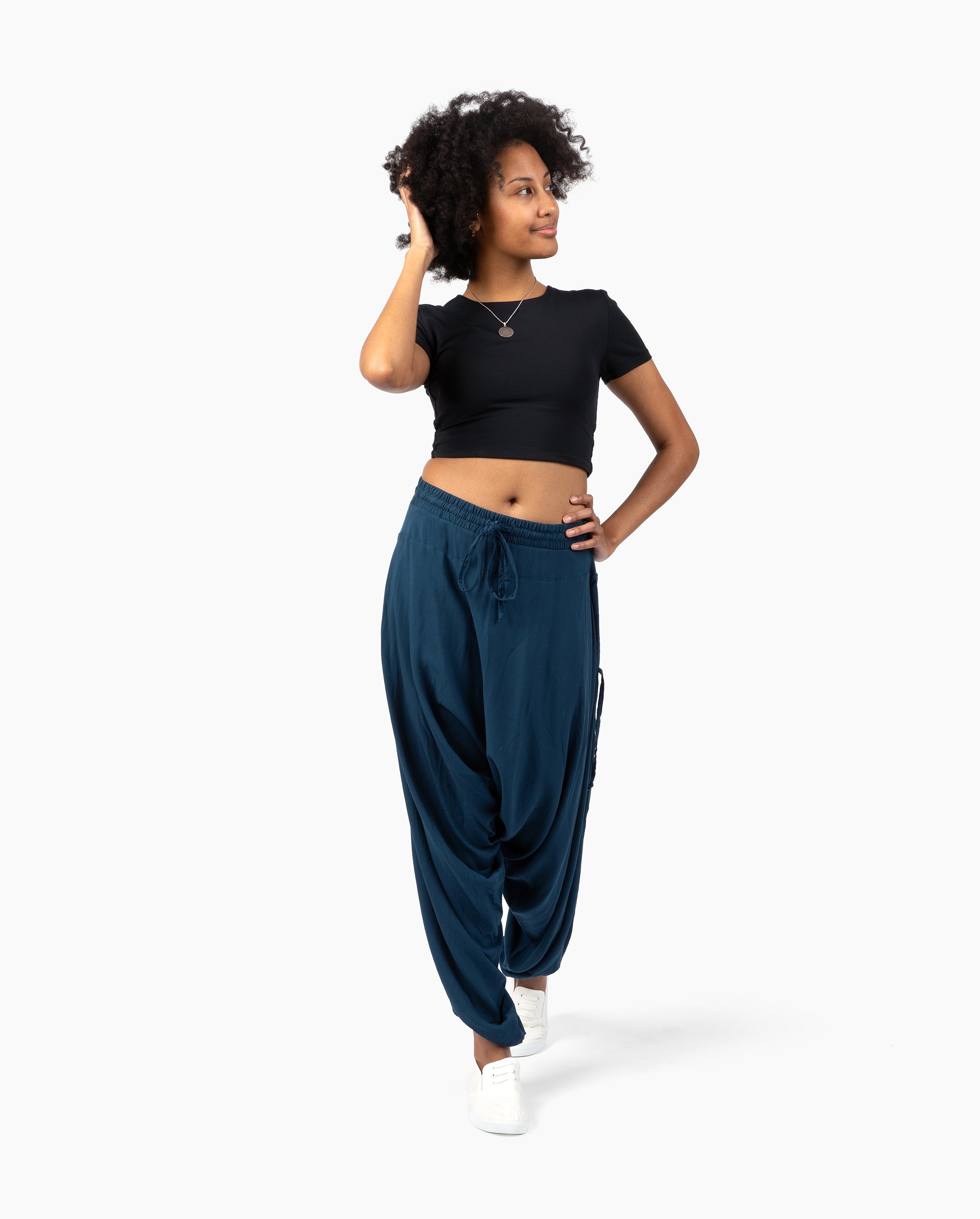 A woman modeling a black short-sleeve crop top and dark teal blue harem pants with white sneakers, standing against a light gray studio background. #color_marine